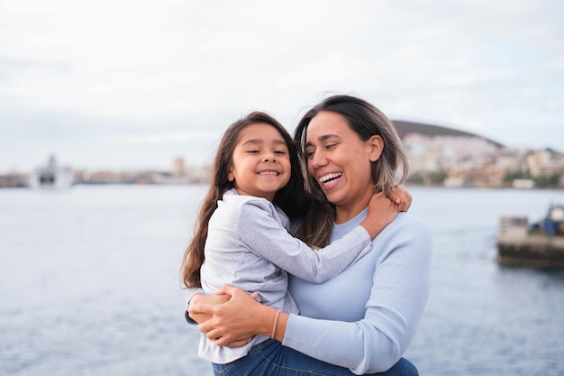 Nahaufnahme der Mutter mit ihrer kleinen Tochter in ihren Armen und Kuscheln am Meer an einem grauen Tag Konzept Mutterschaftslebensstil im Freien