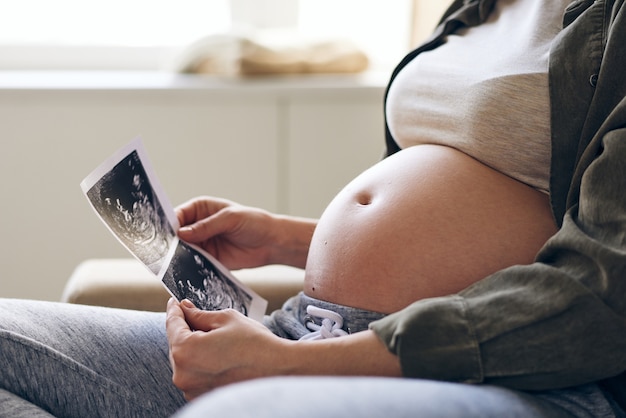 Nahaufnahme der nicht erkennbaren schwangeren frau, die fötales ultraschallbild betrachtet, während sie an baby in ihrem mutterleib denkt