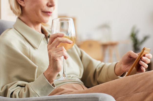 Nahaufnahme der reifen Frau, die auf Sessel mit Weinglas Weißwein sitzt und ihr Handy benutzt