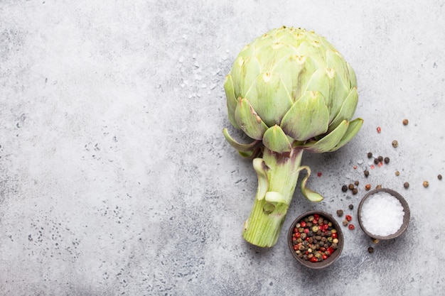 Nahaufnahme der rohen frischen Artischocke mit kochfertigen Gewürzen, ideal als vegetarisches Essen oder Zutat für gesunde Salate und Diäten, rustikaler Hintergrund des grauen Steins