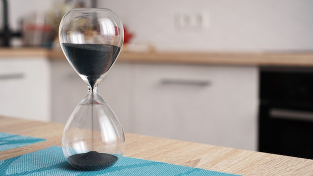 Nahaufnahme der Sanduhr auf einem weißen Holztisch mit defokussierter Küche. Zeitkonzept zum Kochen.