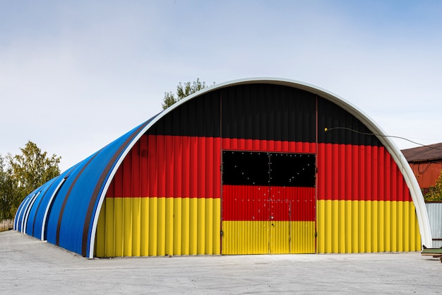 Nahaufnahme der Staatsflagge von Deutschland malte auf der Metallwand eines großen Lagers das geschlossene Gebiet gegen blauen Himmel. Das Konzept der Lagerung von Waren, Eintritt in einen geschlossenen Bereich, Logistik