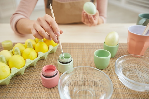 Nahaufnahme der unerkennbaren jungen Frau, die Eier in den Pastellfarben für Ostern malt, während am Tisch in der Küche sitzen, Raum kopieren