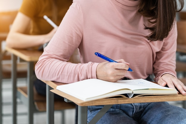 Nahaufnahme der Universitätsstudenten, die im Klassenzimmer Notizen auf dem Notebook schreiben, während sie die Vorlesung hören und studieren. Nahaufnahme von Frauenhänden schreiben auf Notizblock auf Holz-Desktop platziert