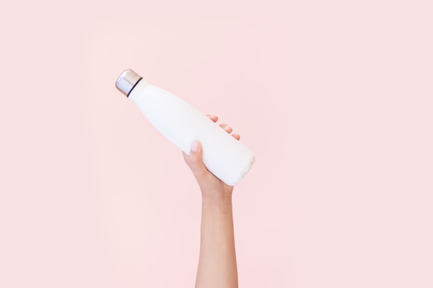 Nahaufnahme der weiblichen Hand, die wiederverwendbare, Stahl-Öko-Thermo-Wasserflasche von Weiß hält, lokalisiert auf Hintergrund der pastellrosa Farbe. Sei plastikfrei. Kein Verlust. Umweltkonzept.