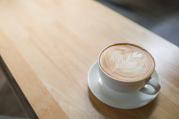 Nahaufnahme der weißen Tasse heißen Kaffeelatte mit Milchschaumherzformkunst auf Holztisch unter Morgensonnenlicht und Schatten- und Kopienraum.