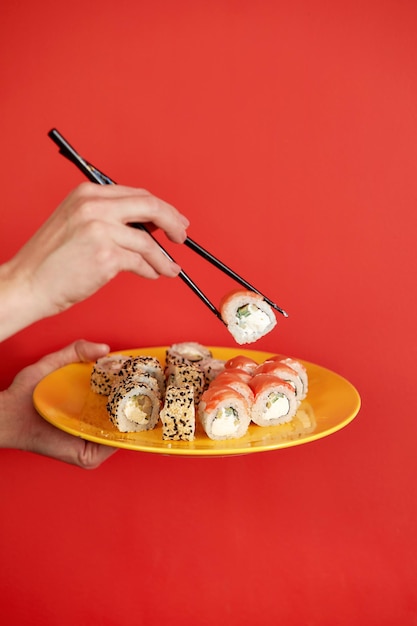 Nahaufnahme des abgeschnittenen Fotos einer weiblichen Hand hält Makizushi-Sushi-Rolle, serviert auf einem Teller traditionelles japanisches Essen, isoliert auf rotem Hintergrund im Studio