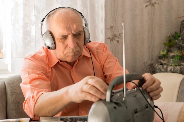 Nahaufnahme des alten Mannes, der Nachrichtensendungen im Radio mit Kopfhörer hört, während er im Wohnbereich sitzt.