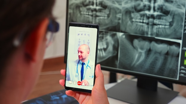 Nahaufnahme des Assistenten, der einen Videoanruf mit einem spezialisierten Stomatologen mit Smartphone hat, der in einer modernen Zahnklinik vor einem PC mit digitalem Röntgen sitzt. Zahnarzt erklärt Patientensymptome