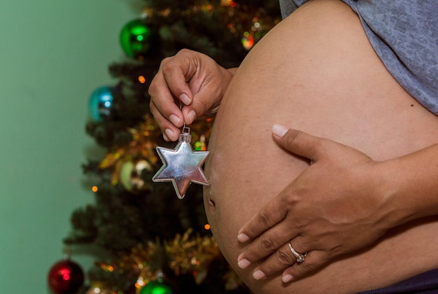 Nahaufnahme des Bauches einer schwangeren Frau, die einen Weihnachtsstern mit dem Weihnachtsbaum im Hintergrund hält