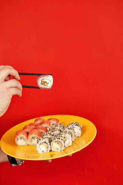 Nahaufnahme des beschnittenen Fotos des männlichen Griffs in den Händen Makizushi-Sushi-Rolle, serviert auf dem Teller traditionelles japanisches Essen isoliert auf rotem Hintergrund im Studio Kopieren Sie Raumwerbung nach oben