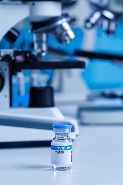 Nahaufnahme des Coronavirus Covid 19-Impfstoffs in einer kleinen Glasfläschchenflasche zum Schutz vor Injektionen auf dem Labortisch vor dem verschwommenen Hintergrund des wissenschaftlichen Mikroskops im Krankenhaus.