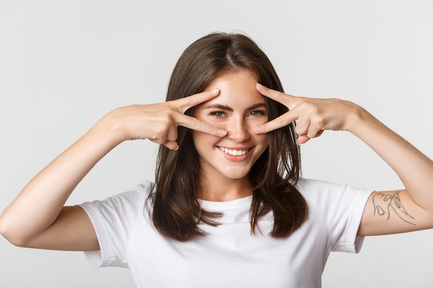 Nahaufnahme des frechen attraktiven brünetten Mädchens, das Friedensgesten über Augen zeigt und glücklich lächelt.