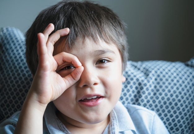 Nahaufnahme des Gesichtes glückliches Kind, das mit lächelndem Gesicht durch die Finger in die Kamera schaut, positives Kind, das Schild mit den Händen zeigt, Schnappschuss von süßen glücklichen Kindern, die Spaß beim Spielen zu Hause haben.