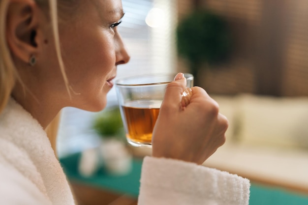 Nahaufnahme des Gesichts einer charmanten, ruhigen jungen Frau im weißen Bademantel, die nach einer Spa-Behandlung einen Schluck aus einer Tasse mit frischem Kräutertee trinkt. Konzept der Körperpflege und Wiederherstellung des spirituellen Friedens