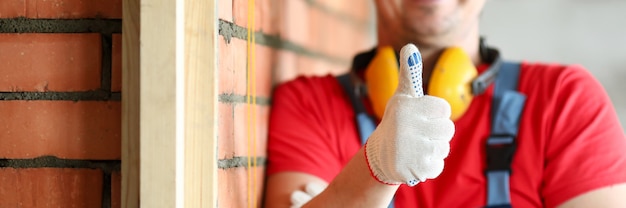 Nahaufnahme des glücklichen männlichen Zimmermanns, der Daumen oben zeigt. Lächelnder Baustellenarbeiter, der am Projekt arbeitet. Rote Backsteinmauer. Renovierungs- und professionelles Baukonzept