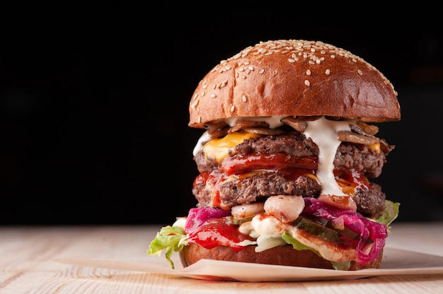Nahaufnahme des großen saftigen doppelten Burgers mit weißer Soße, Käse, Ketchup, Gurken, Pilze und rote Zwiebel auf schwarzem Hintergrund mit Kopienraum. Selektiver Fokus.