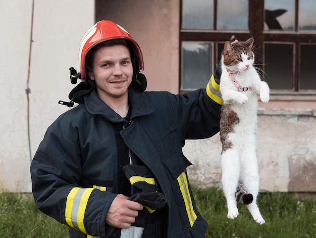 Nahaufnahme des heldenhaften Feuerwehrmanns in Schutzanzug und rotem Helm hält gerettete Katze in seinen Armen. Feuerwehrmann im Löscheinsatz. Foto in hoher Qualität