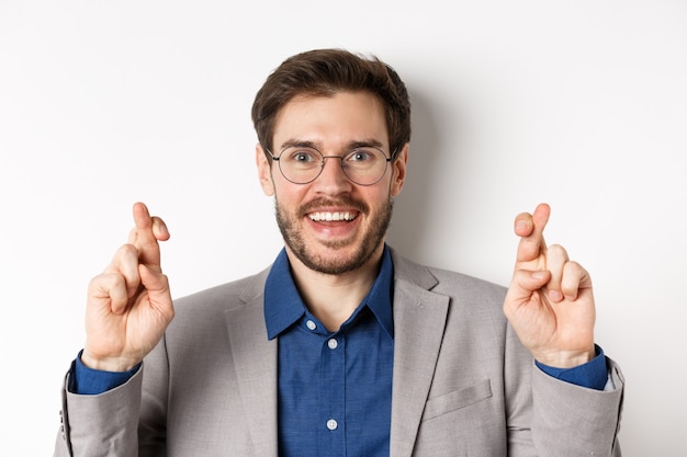 Nahaufnahme des hoffnungsvollen männlichen Unternehmers in Brille und Anzugkreuzfinger für viel Glück, Lächeln und Wunsch machen, für gute Ergebnisse beten, weißer Hintergrund.