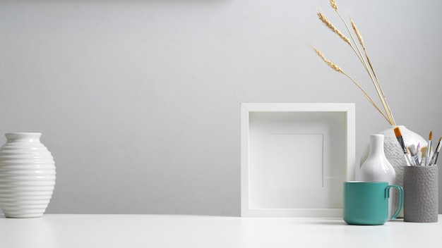 Nahaufnahme des Home-Office-Schreibtisches mit Kopierraum, Modellrahmen, Pinseln und Dekorationen im weißen Konzept