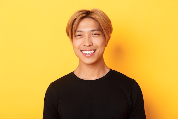Nahaufnahme des hübschen stilvollen asiatischen Kerls mit hellem Haar, glücklich über gelbem Hintergrund lächelnd