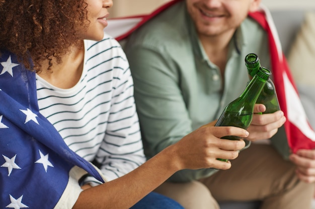 Nahaufnahme des jungen Paares der gemischten Rasse, das Bierflaschen klirrt, während es amerikanische Flagge trägt