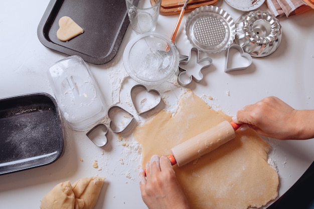 Nahaufnahme des Kochvorgangs. Junge weibliche Hände halten Küchenrolle und rollen damit den Teig auf dem Tisch aus. Teig zum Backen zu Hause herstellen. Küchengeräte sind in der Nähe. Viele Keksformen.