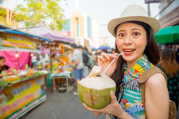 Nahaufnahme des lächelnden schönen Frauentouristenporträts kaufen Sie köstliches Kokosnusswasser auf Reiseferienmarkt im Freien mit allen unscharfen Ständen Hintergrund.