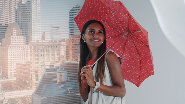 Nahaufnahme des lächelnden schwarzen Modells, das mit rotem Regenschirm auf Barhochstuhl für Fotoshooting des Modemagazins posiert