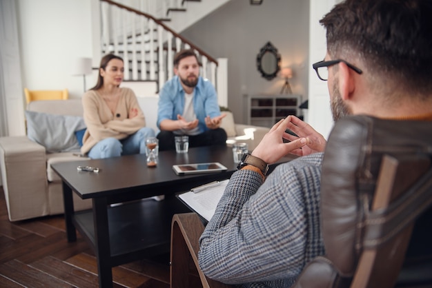 Nahaufnahme des männlichen Psychologen sitzt am Stuhl und hört niedergedrücktes junges Paar am stilvollen gemütlichen Büro. Selektiver Fokus.