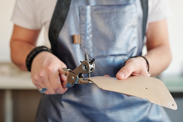 Nahaufnahme des nicht erkennbaren arbeiters in der schürze, die löcher in lederstück unter verwendung des drehwerkzeugs stanzt