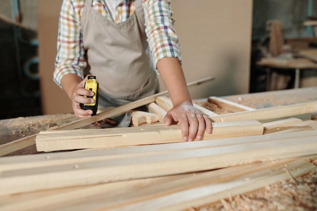 Foto nahaufnahme des nicht erkennbaren jungen in der schürze unter verwendung des maßbandes während der arbeit mit holzdetails in der tischlerei