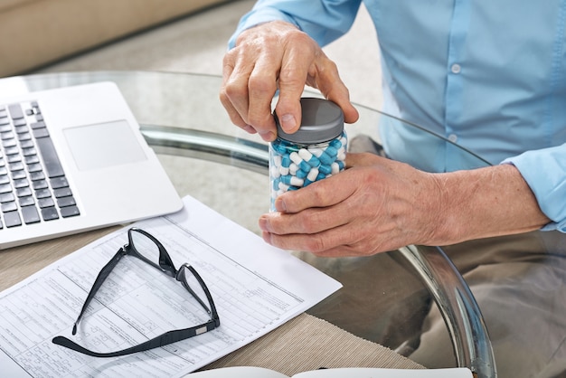 Nahaufnahme des nicht wiedererkennbaren älteren Mannes, der am Tisch mit Laptop und dem Öffnen des Pillenglases sitzt, während er bereit ist, Medikamente zu nehmen