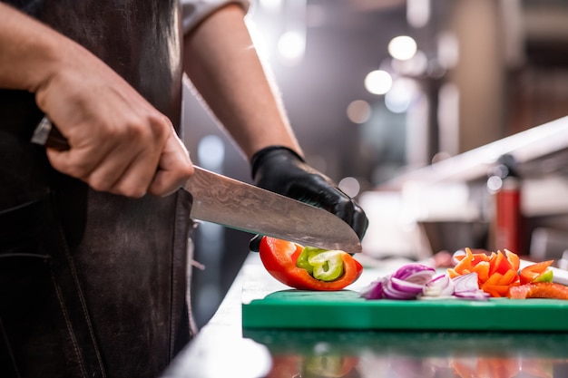 Nahaufnahme des nicht wiedererkennbaren Kochs in der Schürze, die Paprika mit Küchenmesser schneidet, während erfrischender Sommersalat macht