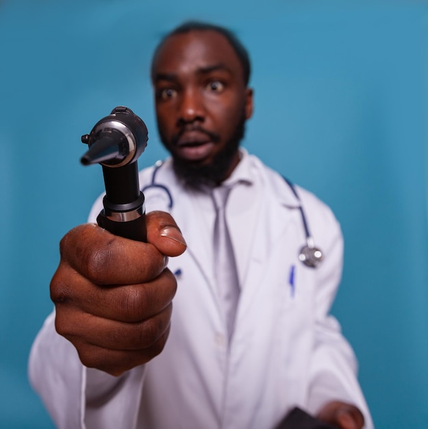 Nahaufnahme des Otoskops in der Hand des afroamerikanischen Arztes mit lustigem Gesichtsausdruck im weißen Laborkittel mit Stethoskop. Nahansicht Sanitäter, der albern hält des HNO-Kits.