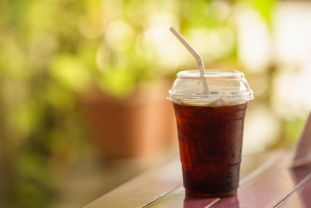 Nahaufnahme des Plastikbechers zum Mitnehmen mit schwarzem Eiskaffee Americano auf Holztisch mit grünem Naturhintergrund