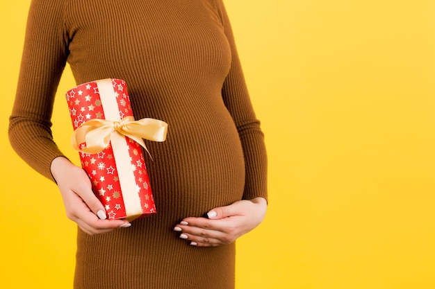 Nahaufnahme des Porträts der schwangeren Frau im braunen Kleid, das eine Geschenkbox auf gelbem Hintergrund hält. Schwangerschaftsfeier. Platz kopieren.