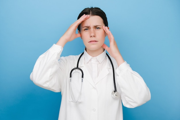 Foto nahaufnahme des porträts einer frustrierten, unglücklichen, traurigen jungen arztfrau in weißer uniform und stethoskop, die isoliert über der blauen studiohintergrundwand posiert. doc ist müde, gelangweilt von der arbeit, enttäuscht, hilflos