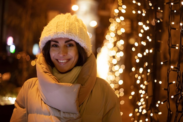 Nahaufnahme des Porträts einer schönen lächelnden Frau mit weißer Wintermütze und Jacke, während sie im Freien mit Weihnachtsbeleuchtung der Stadt im Hintergrund spazieren geht und in die Kamera schaut