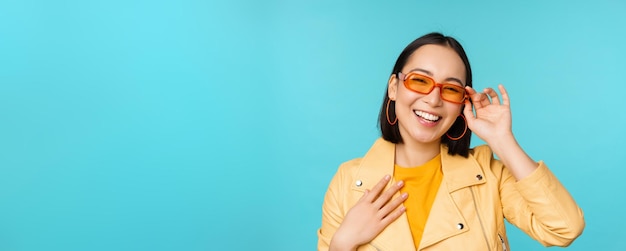 Nahaufnahme des Porträts einer stilvollen asiatischen Frau mit Sonnenbrille, die lacht und lächelt und glücklich aussieht, wenn sie in trendiger Kleidung auf blauem Hintergrund posiert