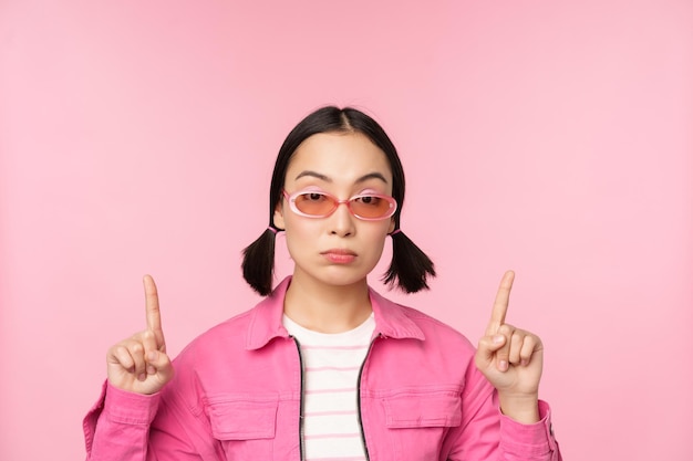 Nahaufnahme des Porträts eines asiatischen, stilvollen Mädchens, das skeptisch aussieht und mit dem Finger nach oben zeigt, mit einem unamüsierten Gesicht, das über rosa Hintergrund steht. Kopierbereich