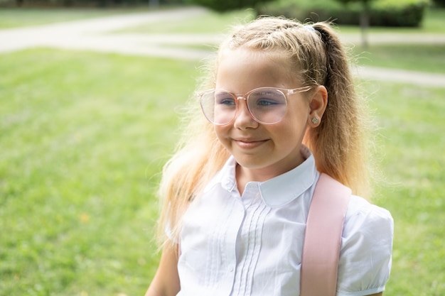 Nahaufnahme des Porträts eines blonden Schulmädchens in einem weißen Hemd mit Brille und einem rosa Rucksack zurück zur Schule