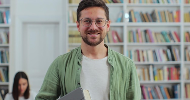 Nahaufnahme des Porträts eines jungen Mannes mit Brille, der ein digitales Tablet im Bücherregal der Bibliothek hält und in die Kamera blickt Hochschulbildungskonzept