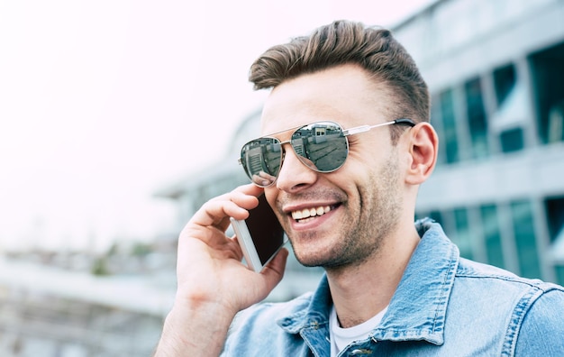 Nahaufnahme des Porträts eines lächelnden, gutaussehenden, trendigen Mannes mit Sonnenbrille und Jeans tragen Gespräche am Telefon im Freien in der Stadt