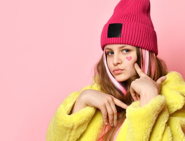 Foto nahaufnahme des porträts eines modernen, selbstbewussten teenager-mädchens auf rosa hintergrund kind mit rosafarbenen haarsträhnen, gekleidet in einen gelben mantel und hut, zeigt auf ein gemaltes herz auf seinem gesicht platz für text