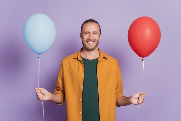 Nahaufnahme des Porträts eines positiven Mannes, der ein Lächeln strahlt und zwei Luftballons an einer lila Wand hält