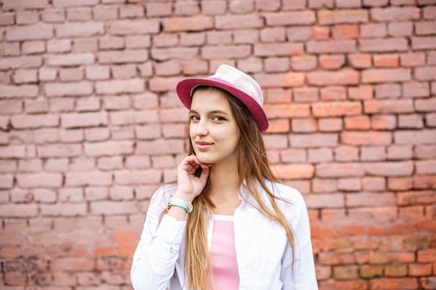 Nahaufnahme des Porträts eines schönen, stilvollen Mädchens mit Hut in der Nähe einer rosa Backsteinmauer als Hintergrund