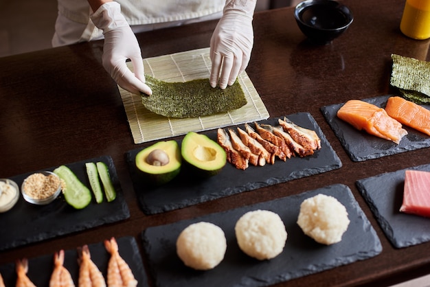 Nahaufnahme des Prozesses der Zubereitung von rollendem Sushi. Die Hände des Küchenchefs halten ein Blatt Nori. Zutaten: Gurke, Lachs, Reis, Avocado auf schwarzen Steinplatten