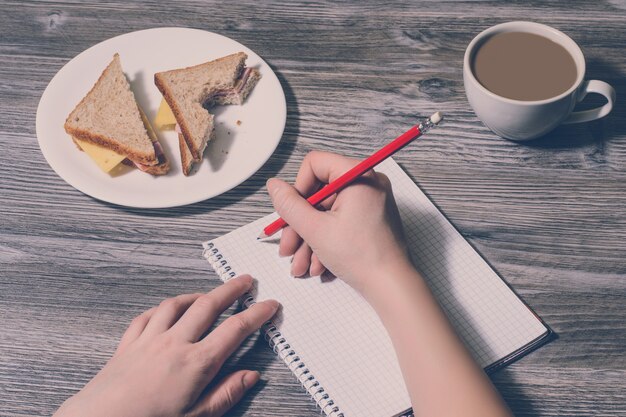 Nahaufnahme des Schreibens von Hand in ein Notizbuch und leckeres Käsesandwich auf einem runden Teller, Tasse Latte. Holzhintergrund, Ansicht von oben