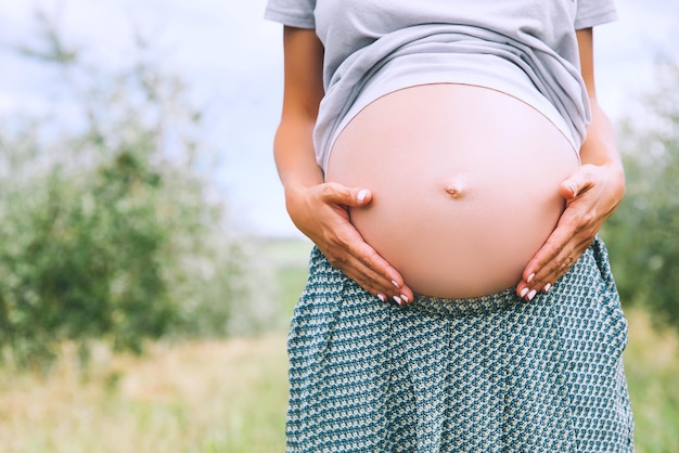 Nahaufnahme des schwangeren Bauches in der Natur im Freien Kopierraum Schwangere Frau hält die Hände auf dem Bauch auf dem natürlichen Hintergrund des grünen Grases Schwangerschaftserwartung Geburt neuer Lebenskonzepte